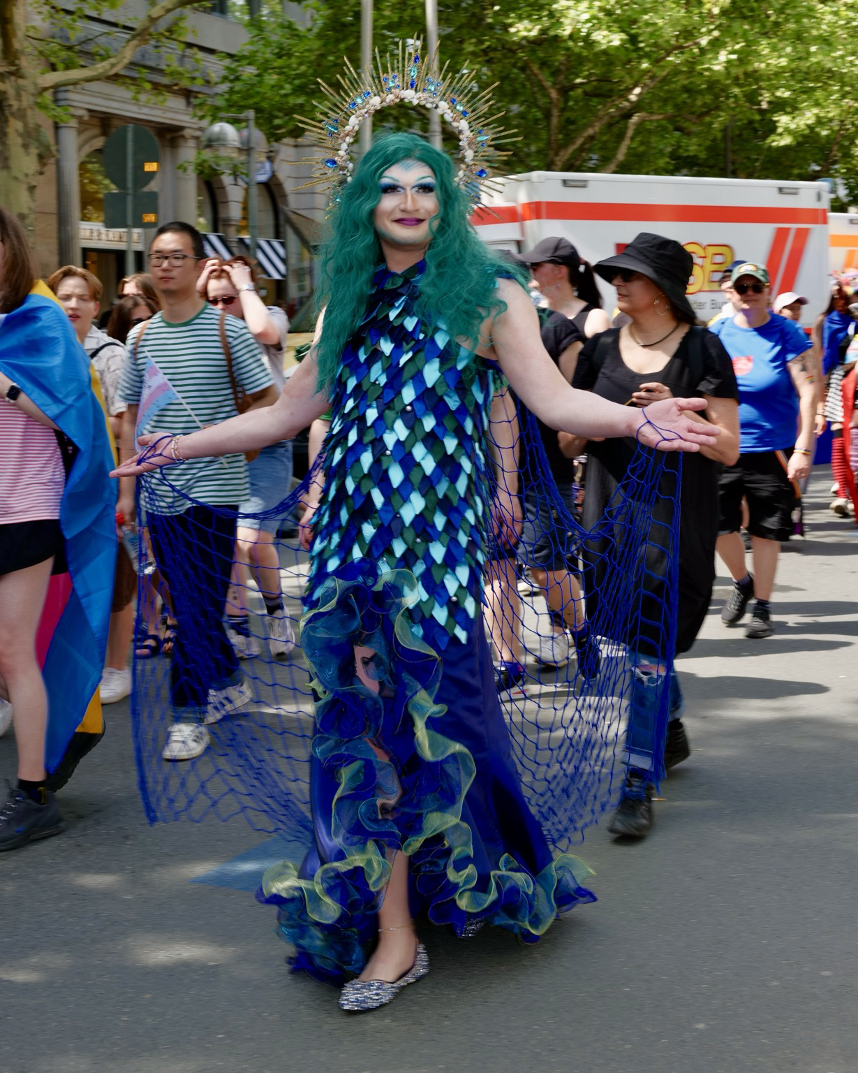 “A person in elaborate drag attire walks in a Pride parade wearing a shimmering blue and teal dress inspired by ocean scales, with flowing netted sleeves and ruffled details. They have long turquoise hair, dramatic makeup, sparkling shoes, and a large halo-like crown decorated with blue jewels and gold rays. Parade participants and spectators surround them on a sunny, tree-lined street.