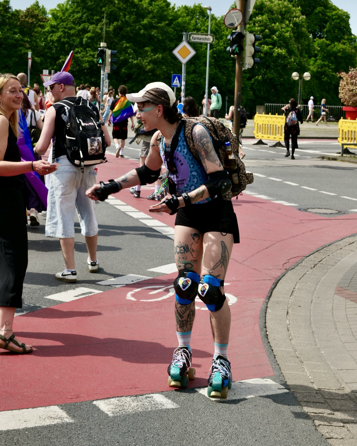 A tattooed person wearing roller skates pauses during a Pride parade to talk with someone at the roadside. They wear protective knee and elbow pads, a sleeveless blue patterned top, black shorts, and a backpack, with visible tattoos on their arms and legs. Other parade attendees and rainbow flags appear in the background along a sunny city street.