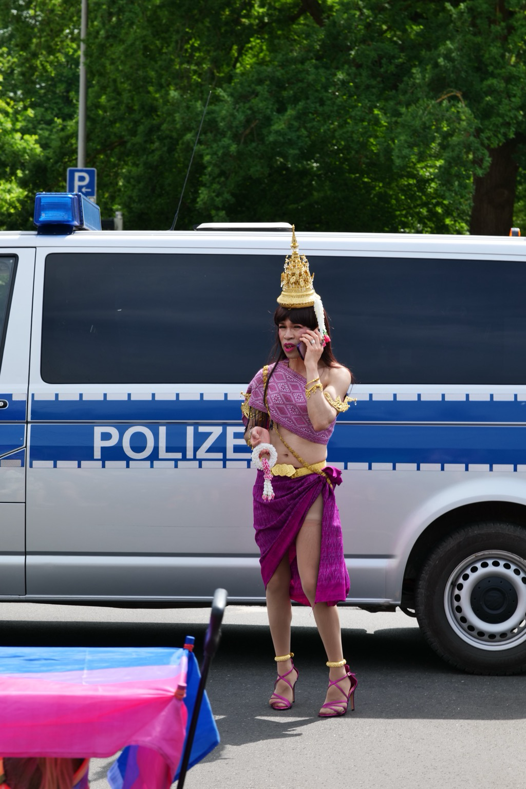 A person wearing ornate traditional-inspired attire stands in front of a German police van while talking on a cellphone during a Pride event. They wear a gold headdress, purple wrap skirt, matching high heels, and gold jewelry. Trees and parade elements are visible in the background on a sunny day.
